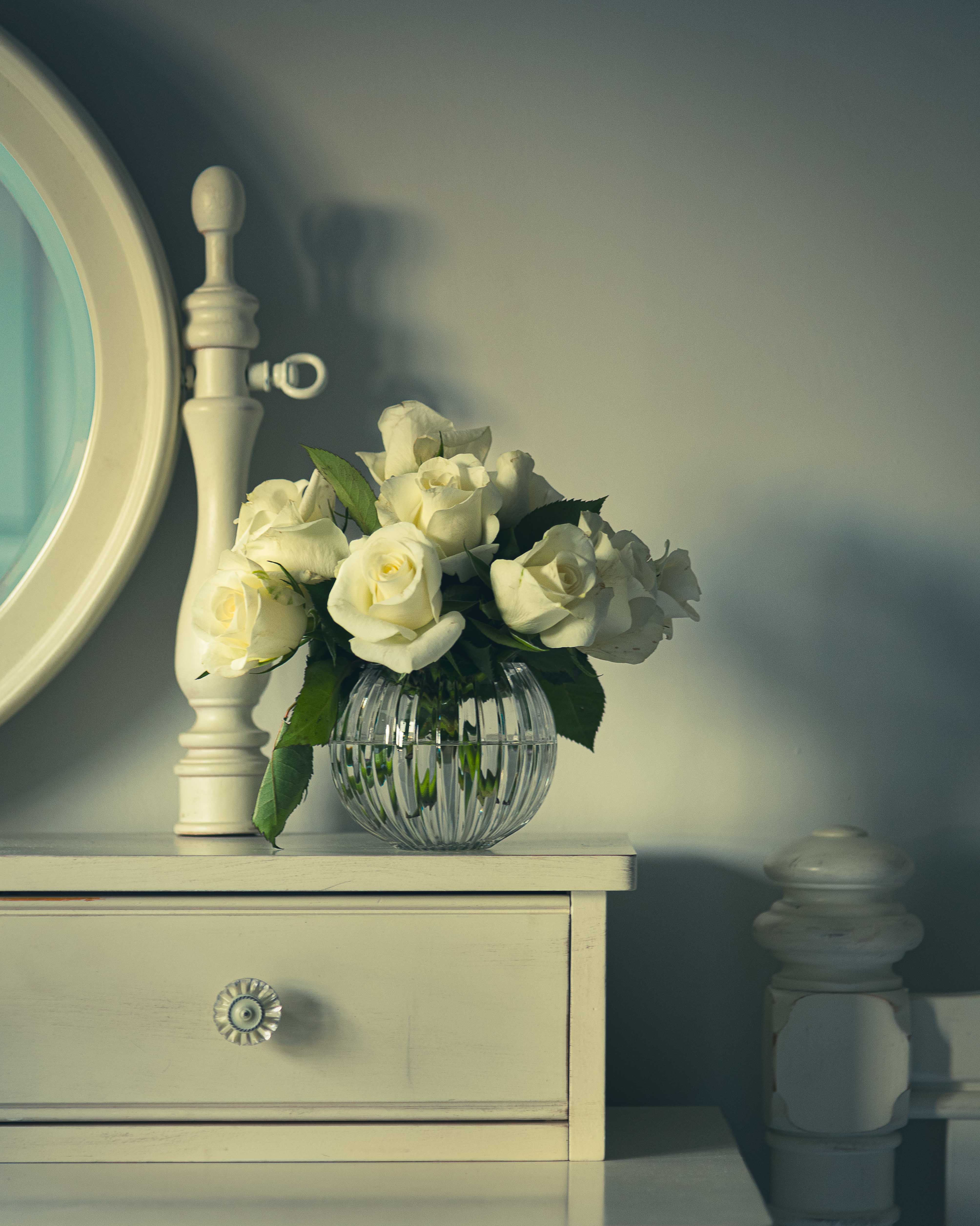 A bouquet of white roses in a clear glass vase on a light-colored dresser next to a round mirror, with soft shadows and a serene atmosphere.