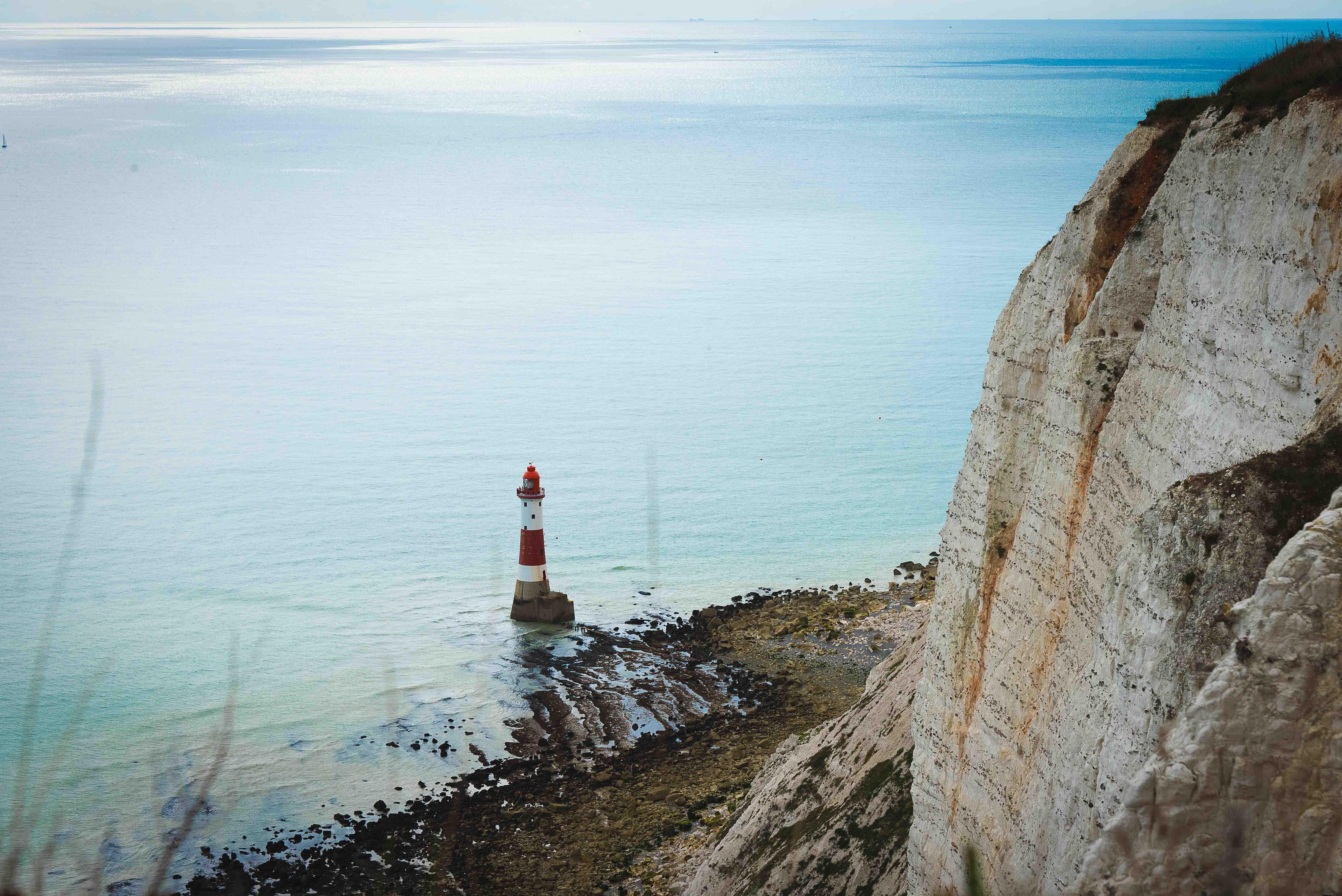Beachy Head Lighthouse stands on a rocky shore with a cliff in the background, overlooking a calm sea under a cloudy sky.