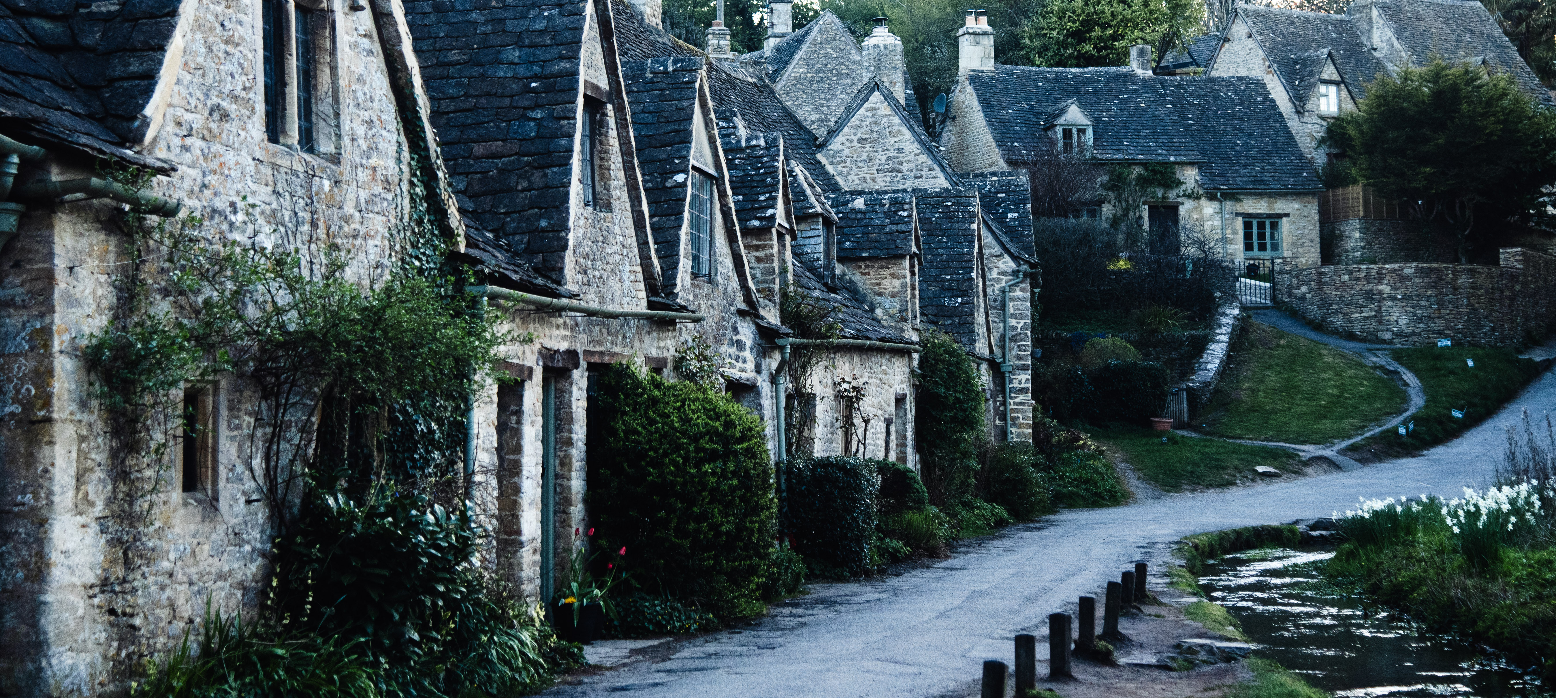 A picturesque view of charming stone cottages lining a quiet, winding road, with lush greenery and blooming flowers alongside a gentle stream.